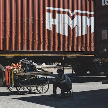 A man sits in shadow next to a cart and HMM cargo container outdoors.