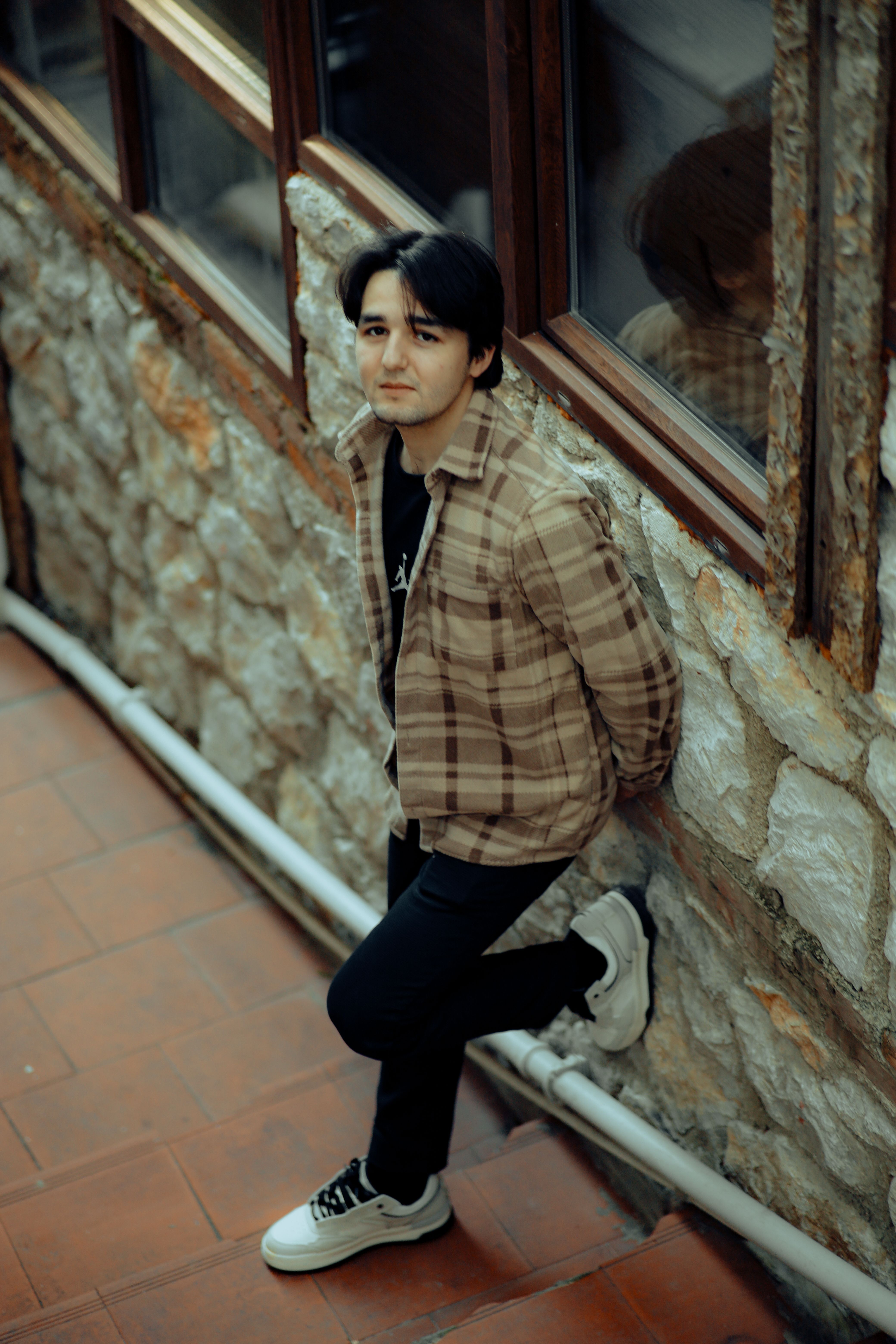 A stylish man leans against a stone wall in an urban setting.