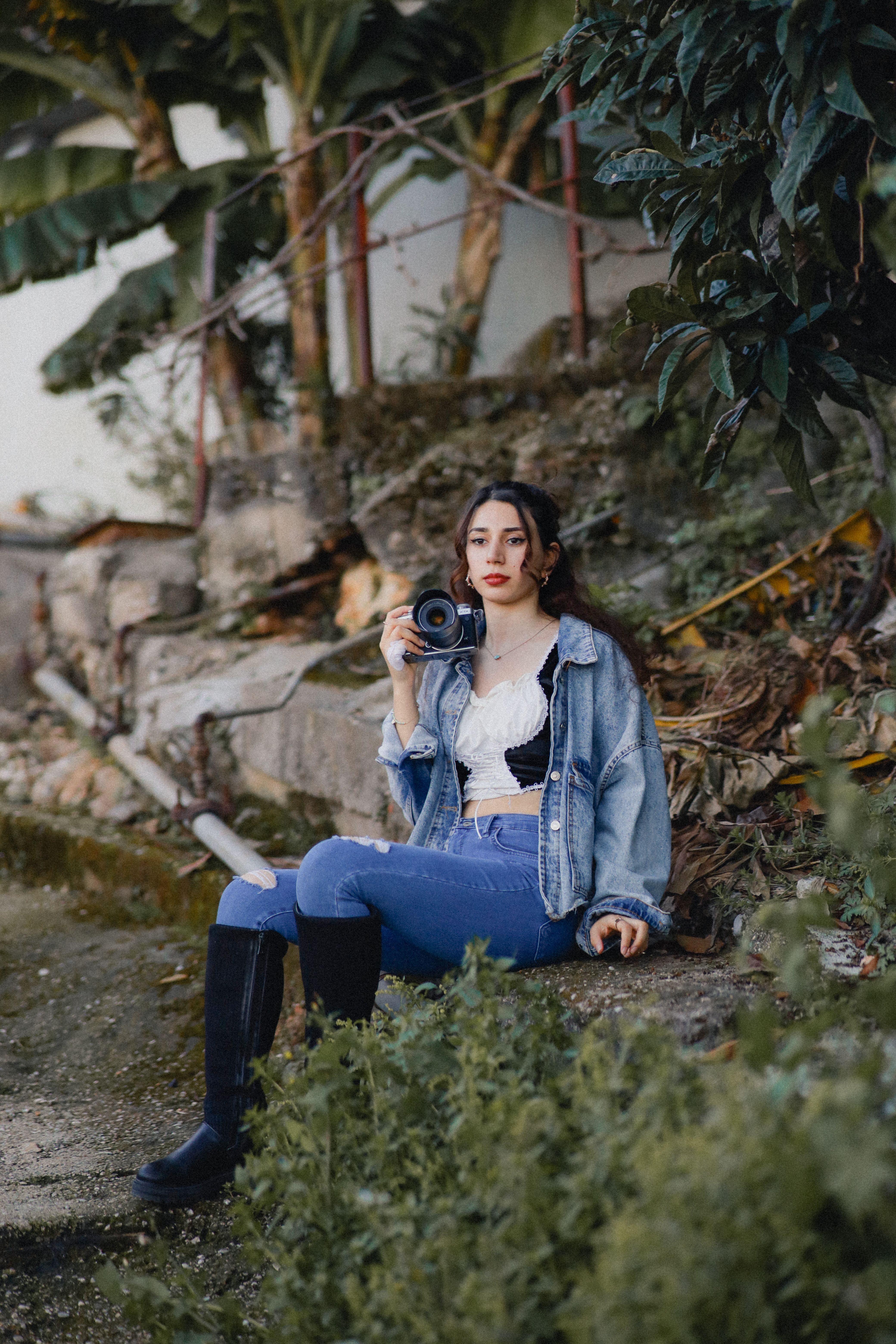 A fashionable young woman in a rustic setting holding a camera, in a stylish denim outfit.