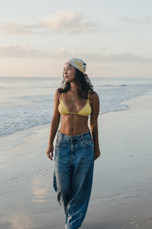 A woman in a yellow bikini and jeans walking on the beach
