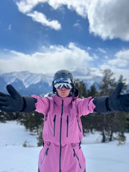 A woman in a pink ski jacket enjoys a snowy mountain day with arms outstretched.