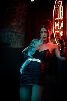 Fashionable woman poses confidently under neon lights, showcasing a chic dress and glamorous style.