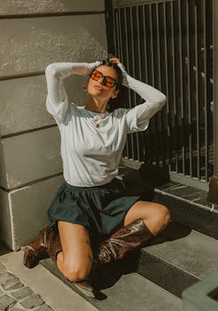 Fashionable woman in sunglasses and boots posing on steps in the sunlight, showcasing a vintage style.