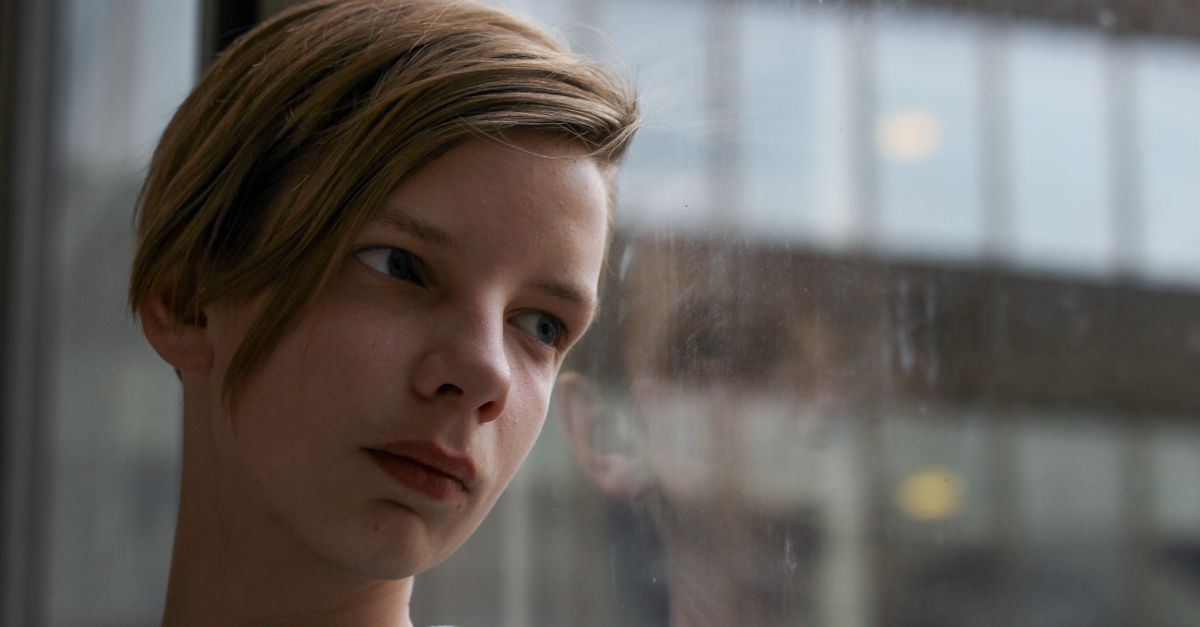 A thoughtful teenager gazing out a window, reflecting in the glass.