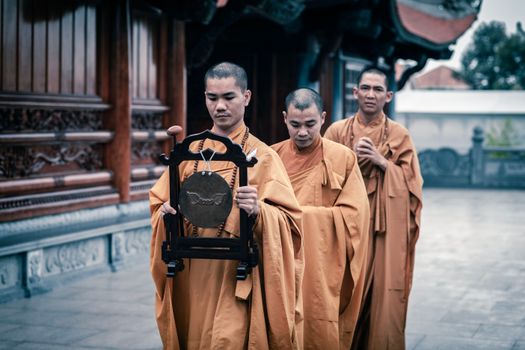 Buddhist monks in traditional robes participate in a temple ceremony holding a ceremonial gong.