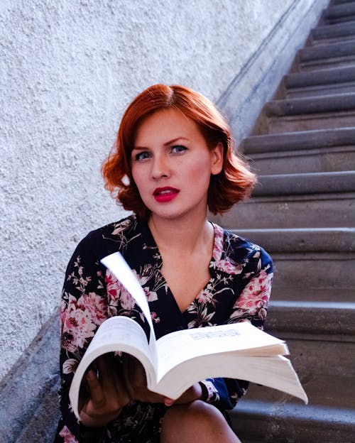 A woman with red hair is sitting on some stairs reading a book