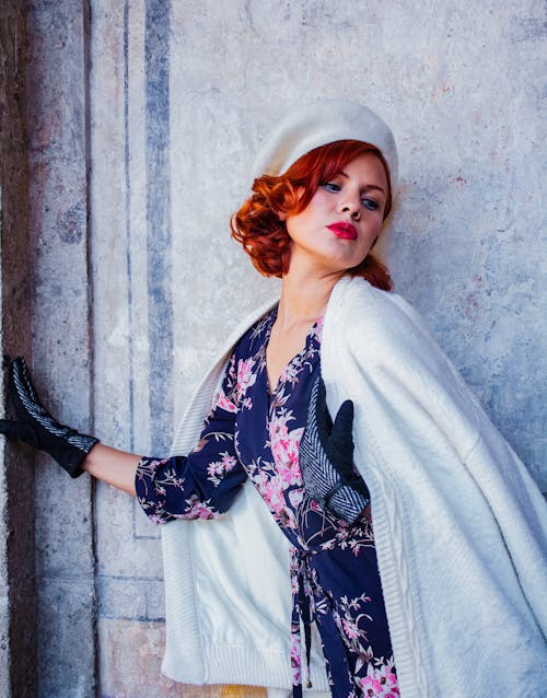 A woman in a white coat and hat leaning against a wall