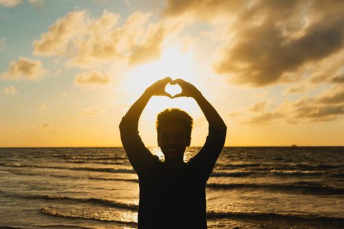 Free Silhouette of a teenager forming a heart shape with raised arms against a vivid seaside sunset. Stock Photo