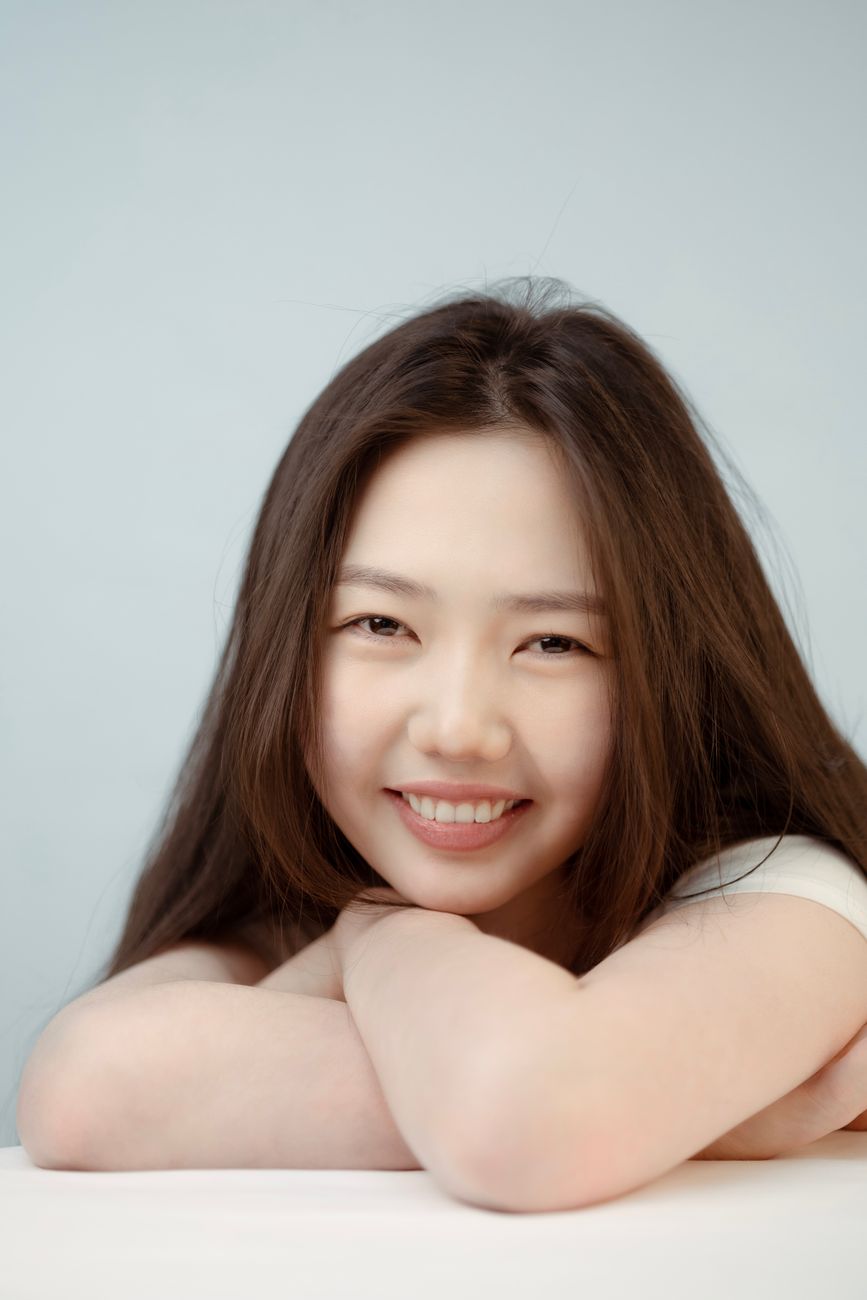 Studio portrait of a smiling woman with long brown hair leaning on a white surface against a white background.