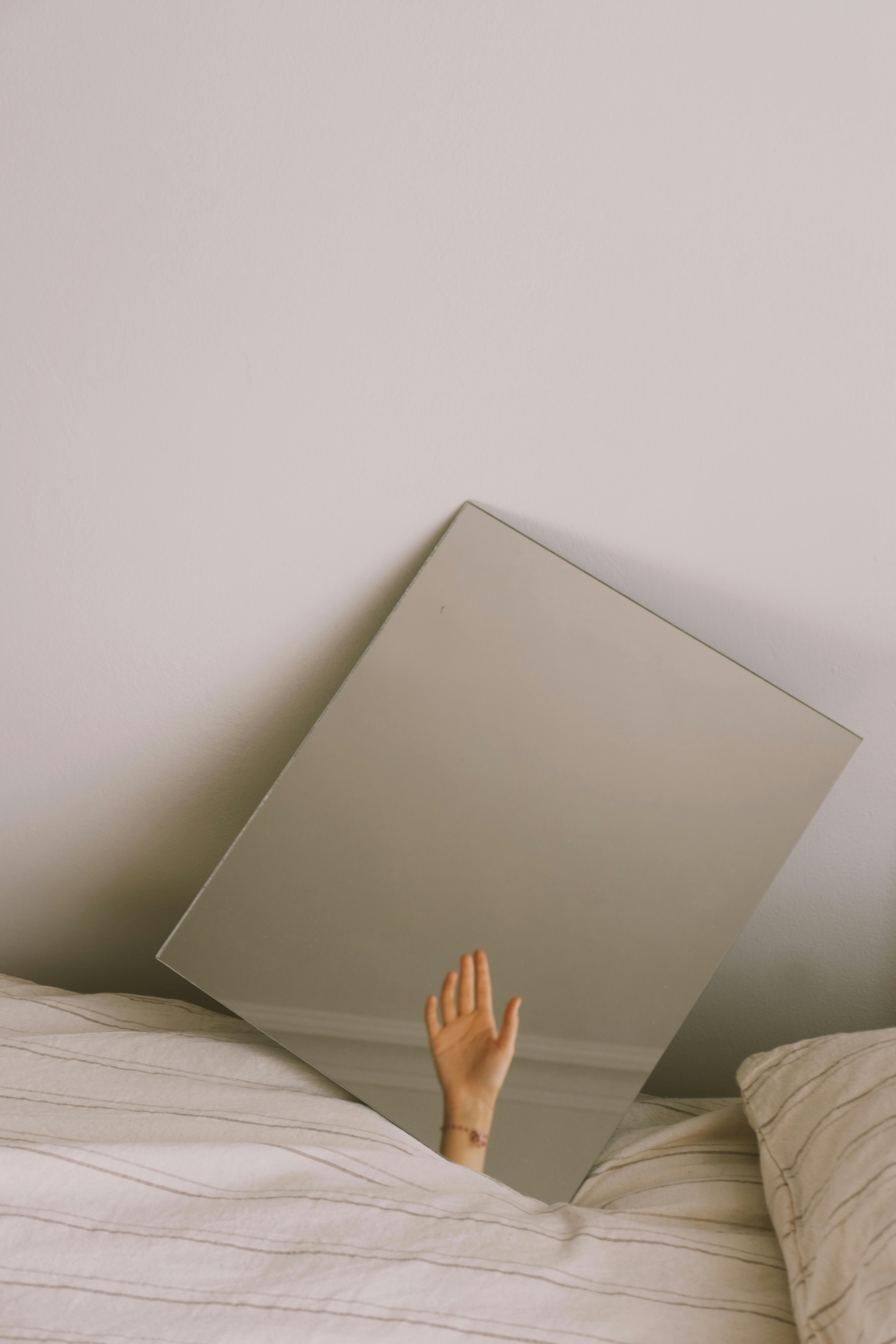 A creative minimalist photo featuring a raised hand reflected in a mirror resting on a bed.