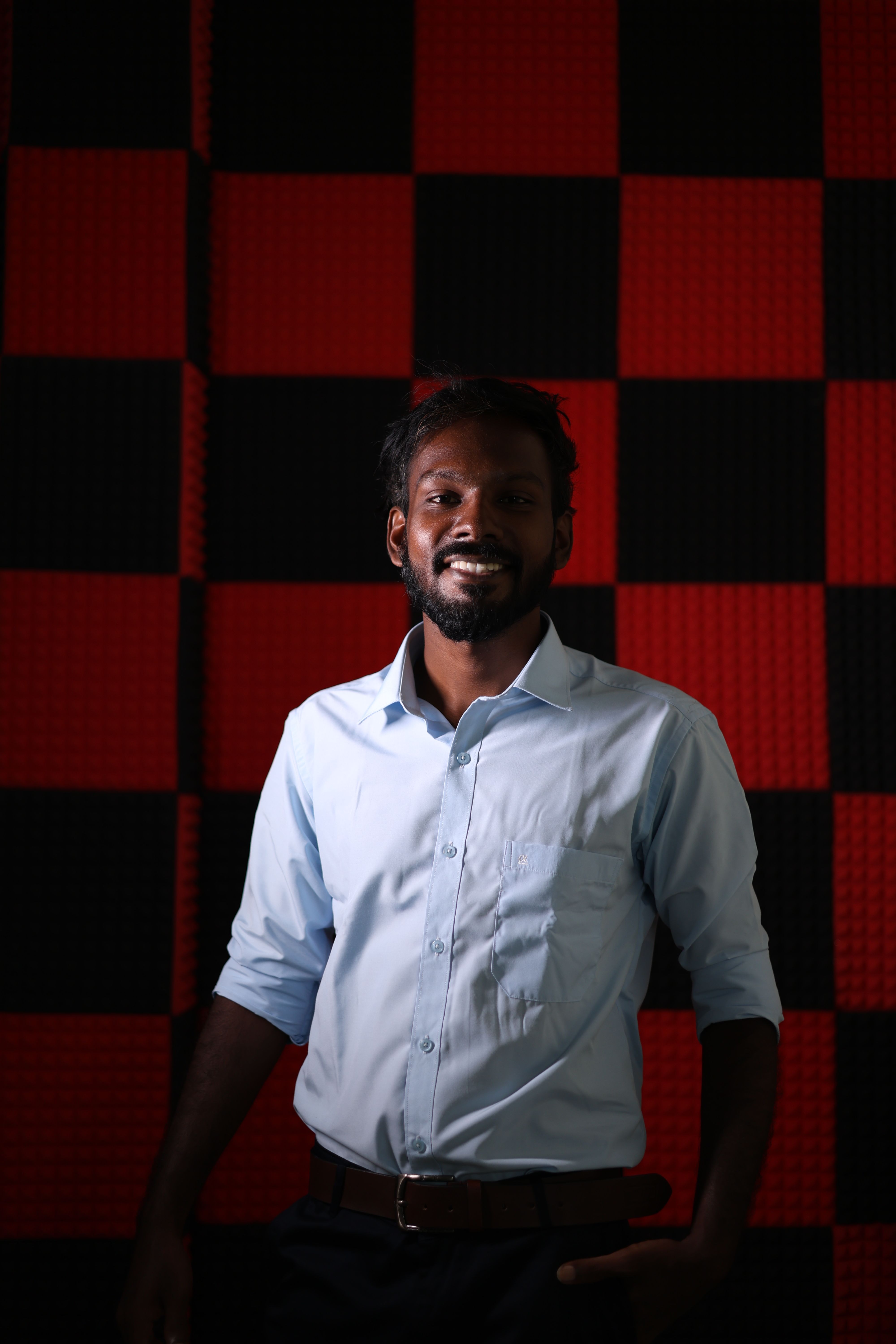Studio portrait of a smiling man in a light blue shirt against a red and black checkered background.