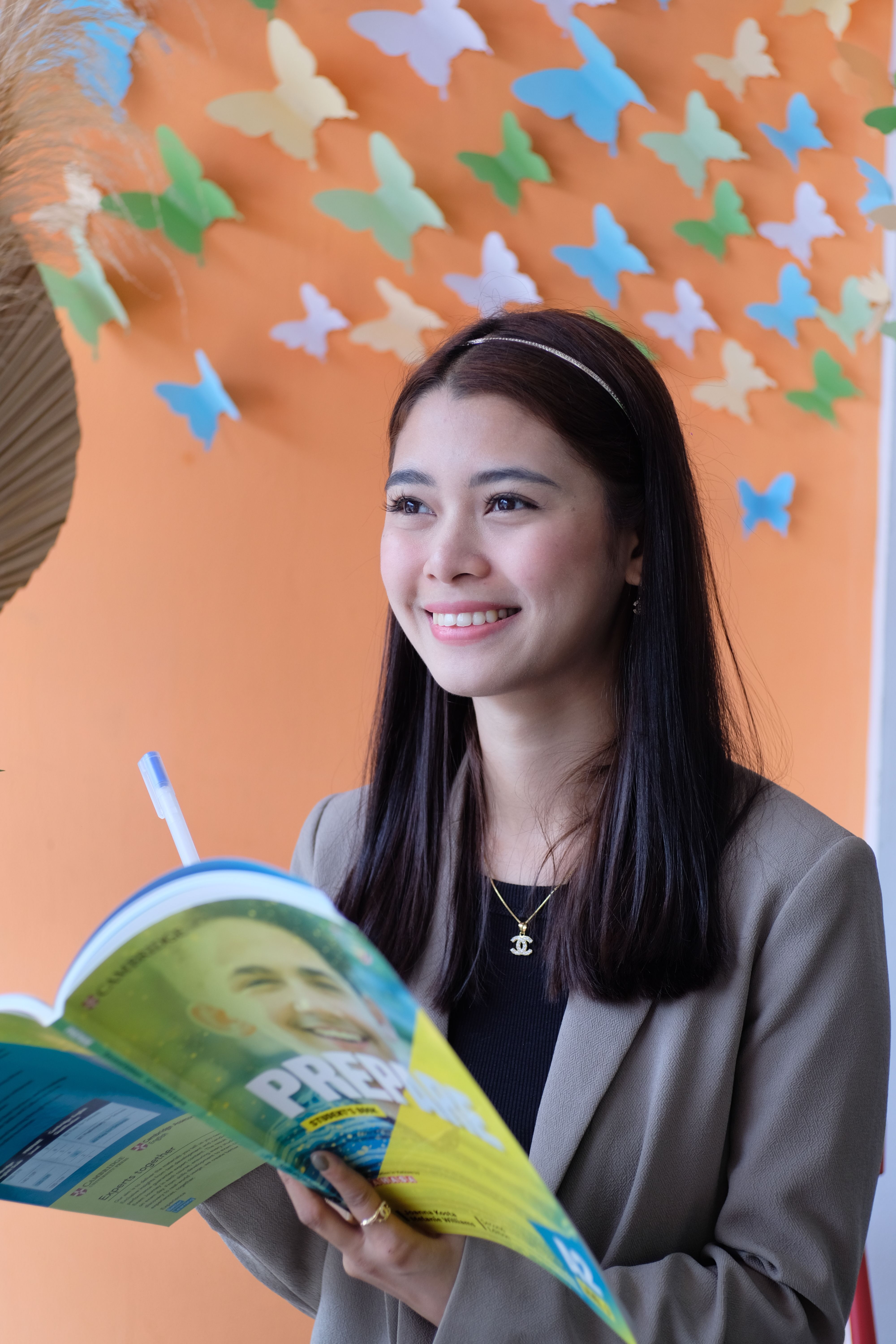 A young woman smiling while holding a book against a colorful butterfly wall background.