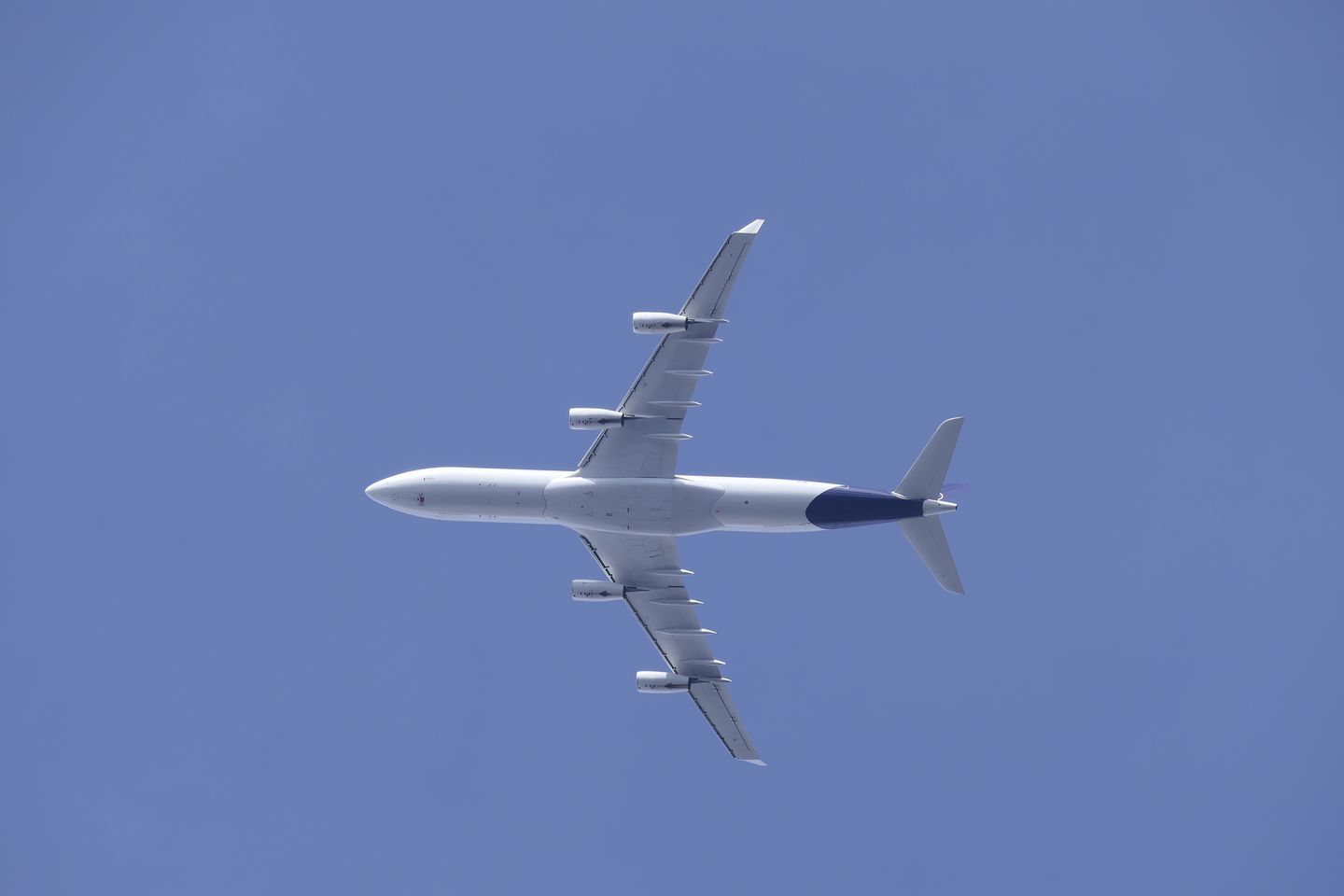 A white four-engine airliner with a dark blue tail, seen from directly below against a clear blue sky