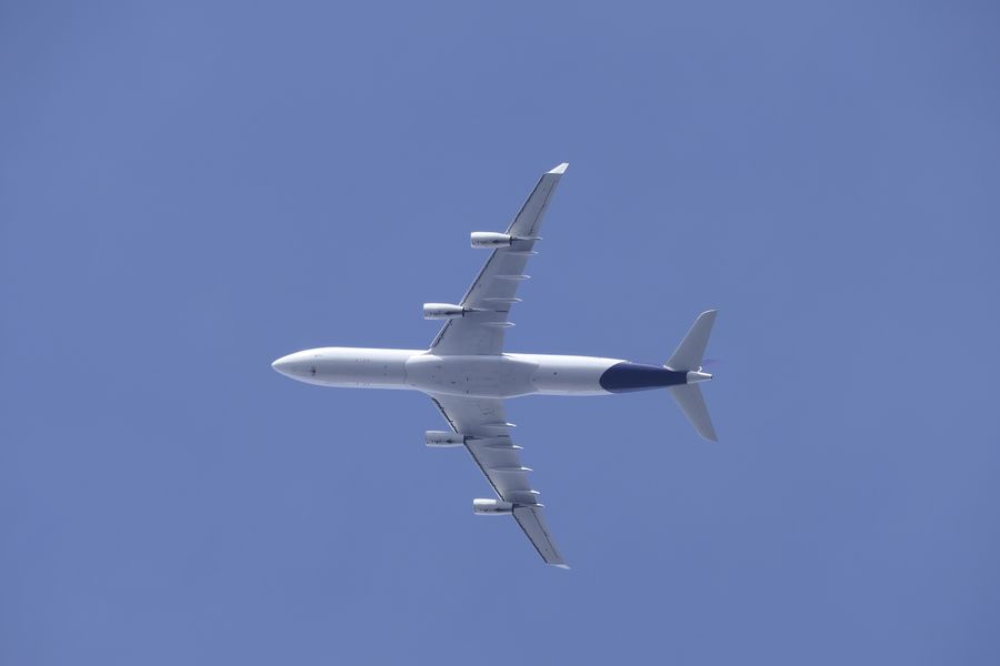 A white four-engine airliner with a dark blue tail, seen from directly below against a clear blue sky