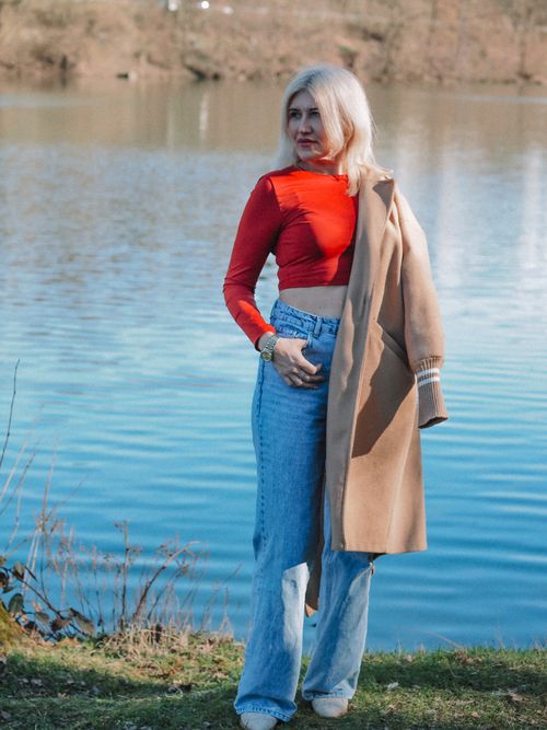 Free Woman standing by a serene lake in a red top and jeans, showcasing autumn fashion. Stock Photo