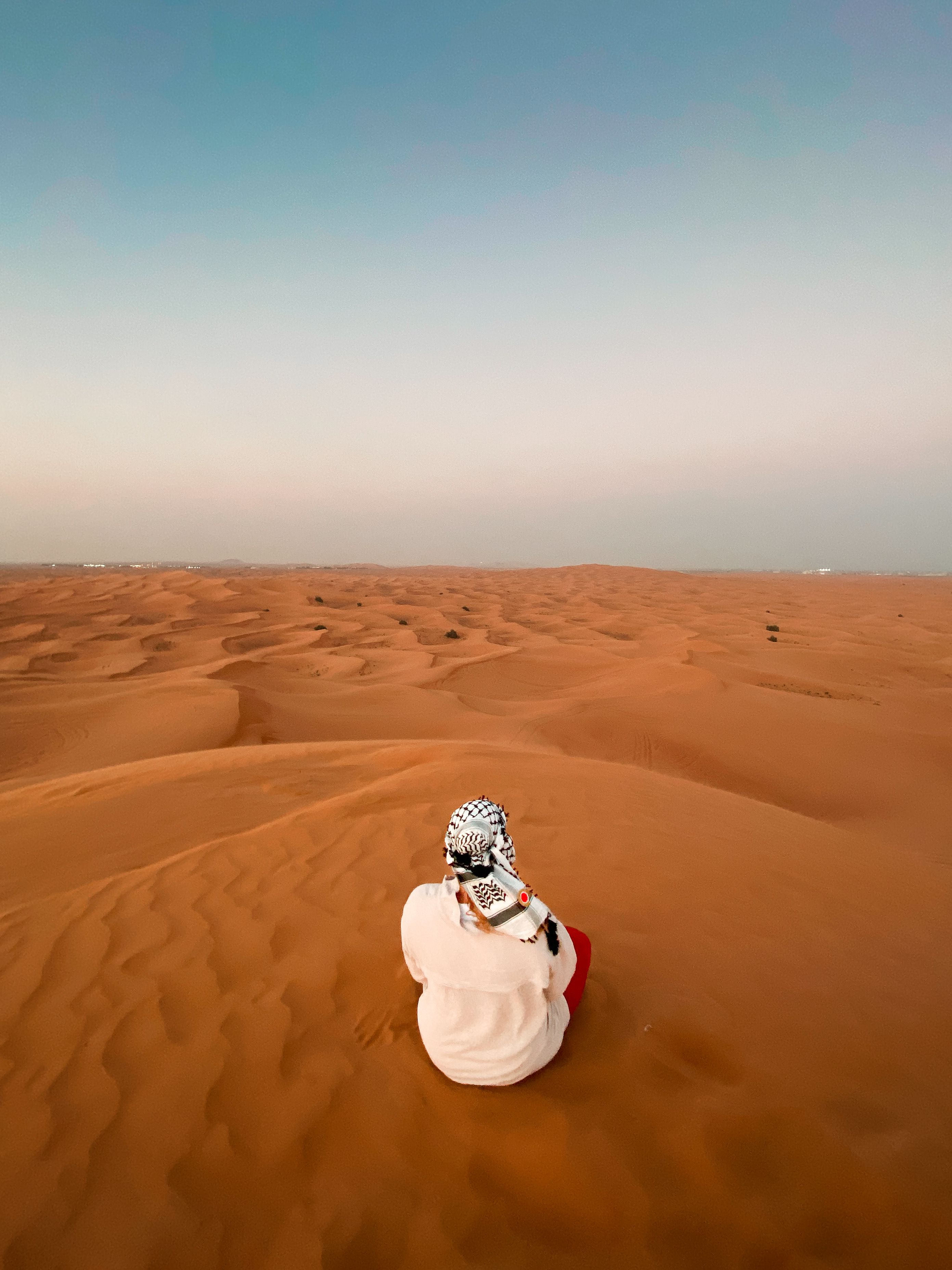 A serene individual sits amidst the vast desert dunes in Dubai under a clear blue sky.