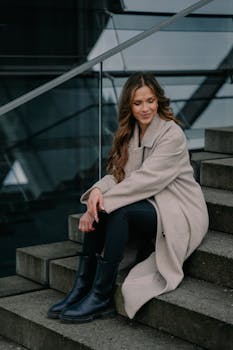 Fashionable woman with long hair sitting on stairs in Berlin, wearing coat and boots, smiling.