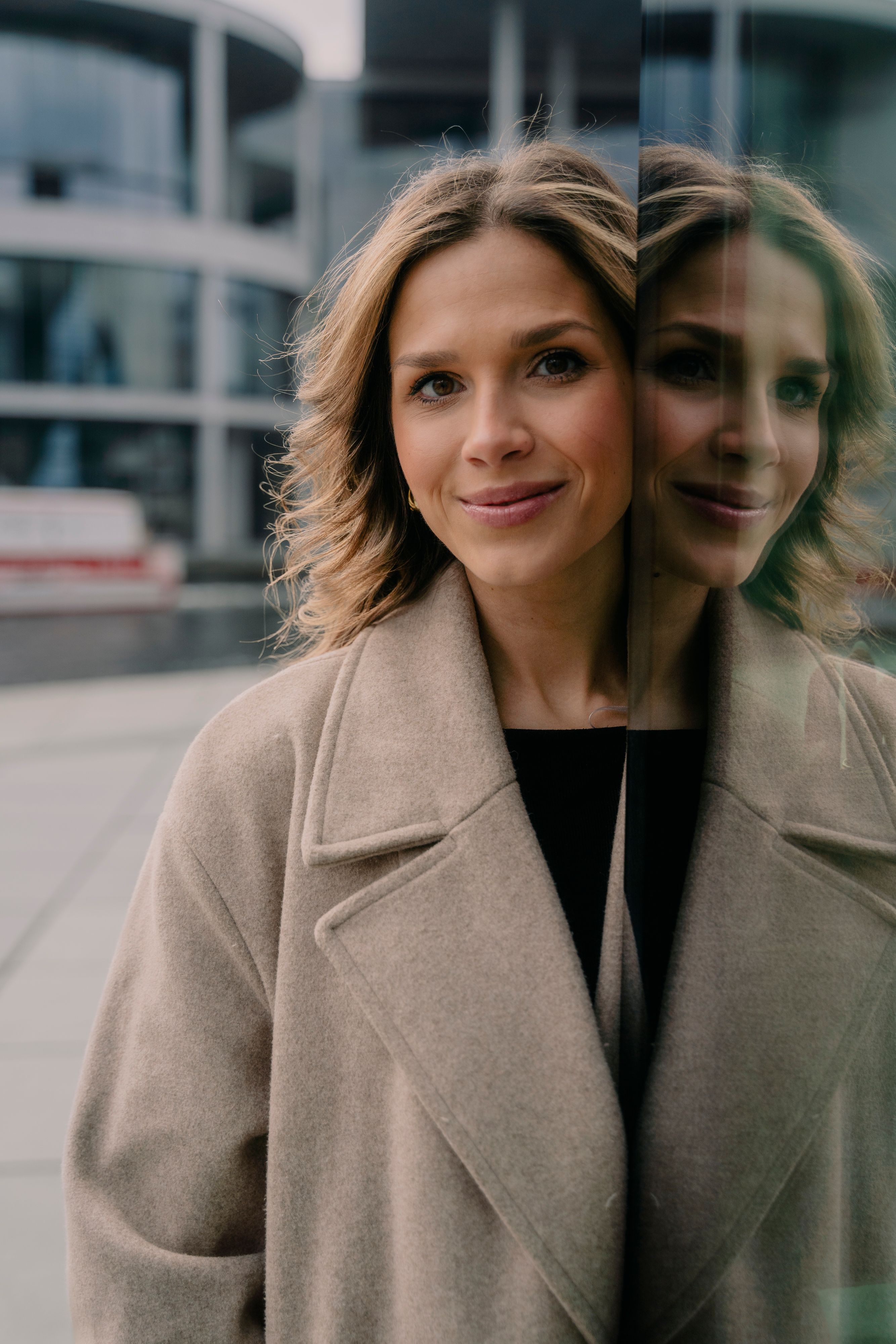 Stylish woman in coat smiles beside modern glass building in Berlin.