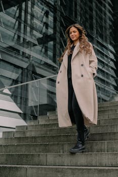 Stylish woman in beige coat walking down stairs in Berlin with modern architecture background.
