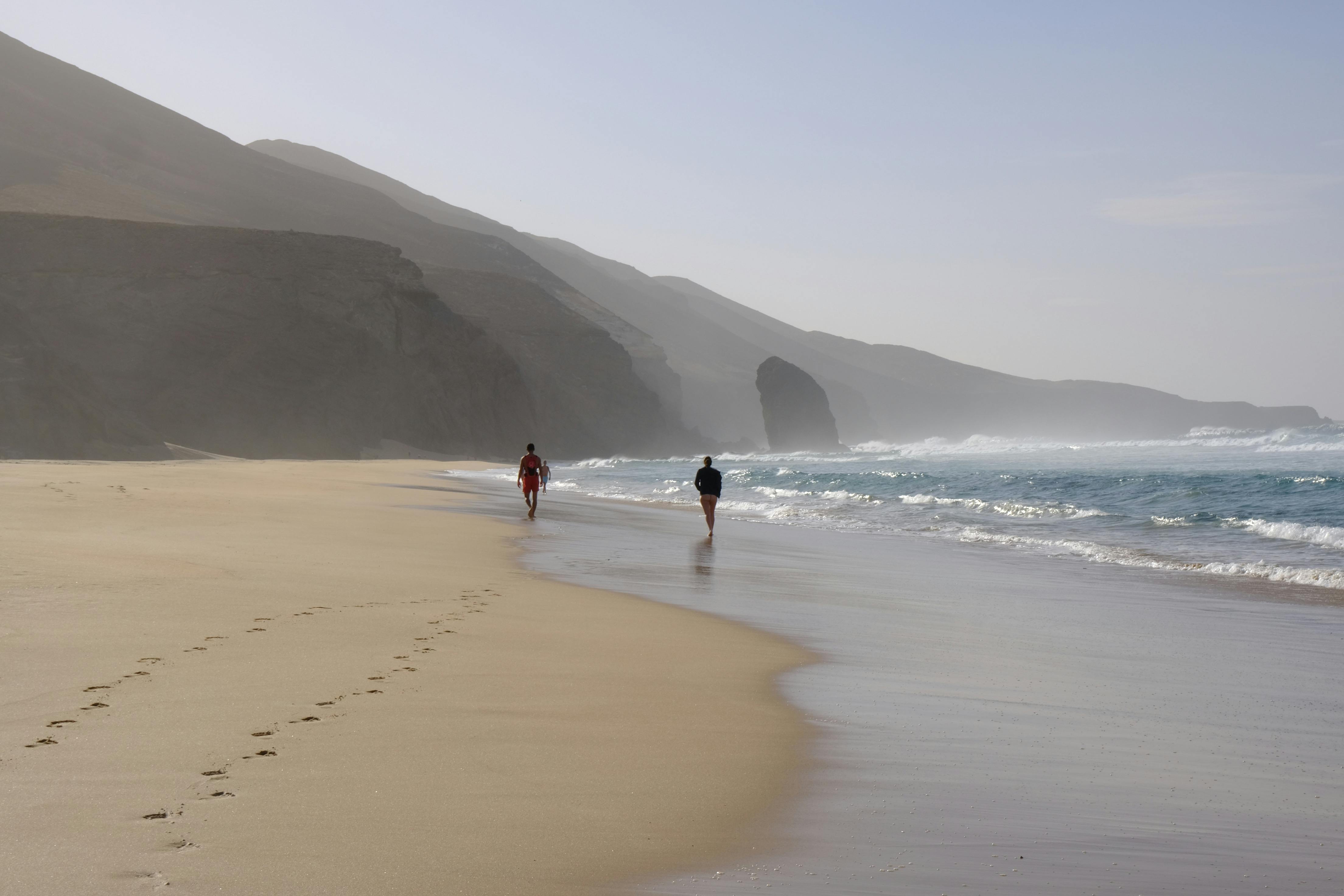 Two people walking along a serene beach with ocean waves and cliffs in a picturesque setting.