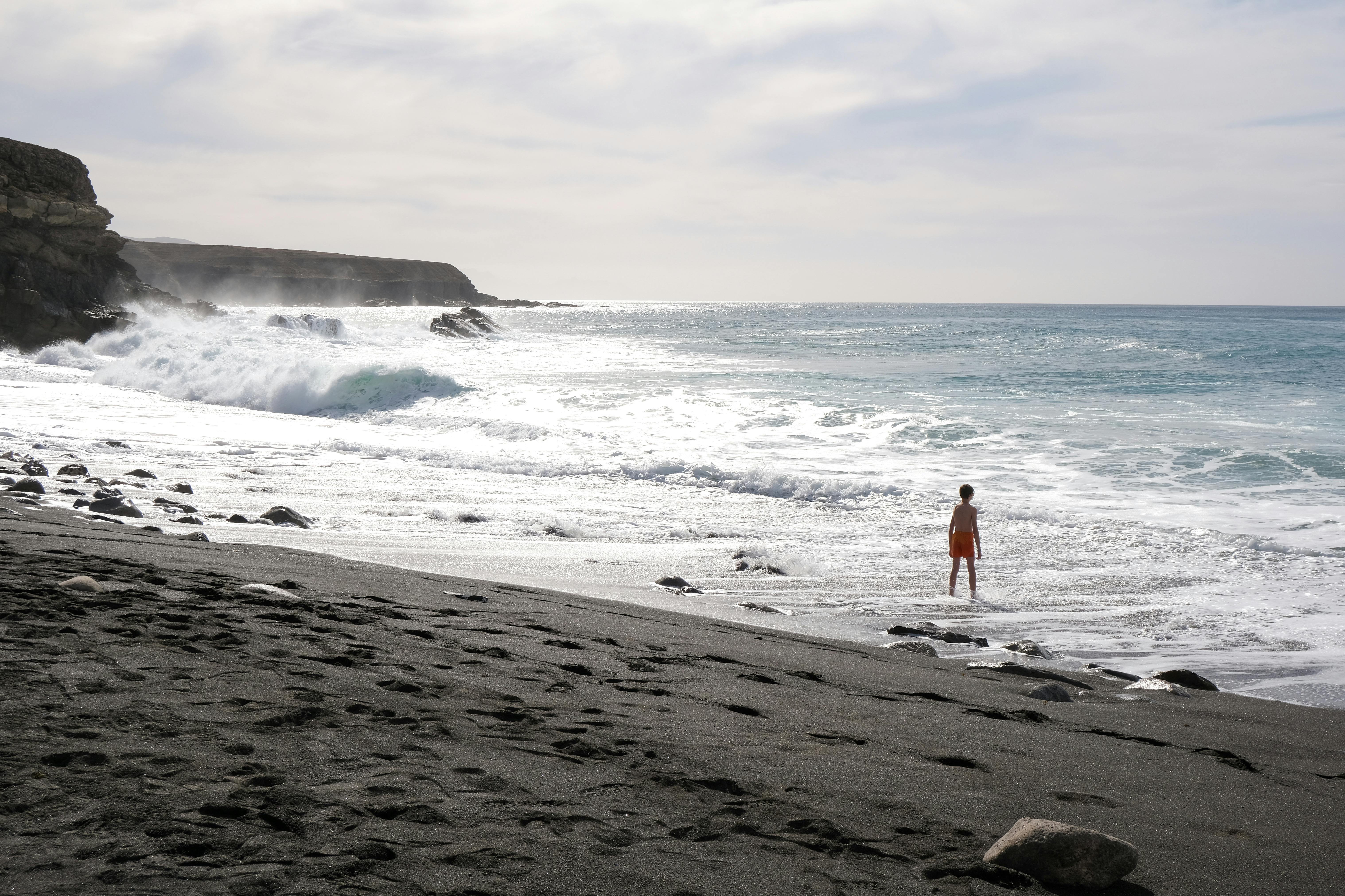 A child stands alone on a rocky beach, watching the waves crash during summer day.