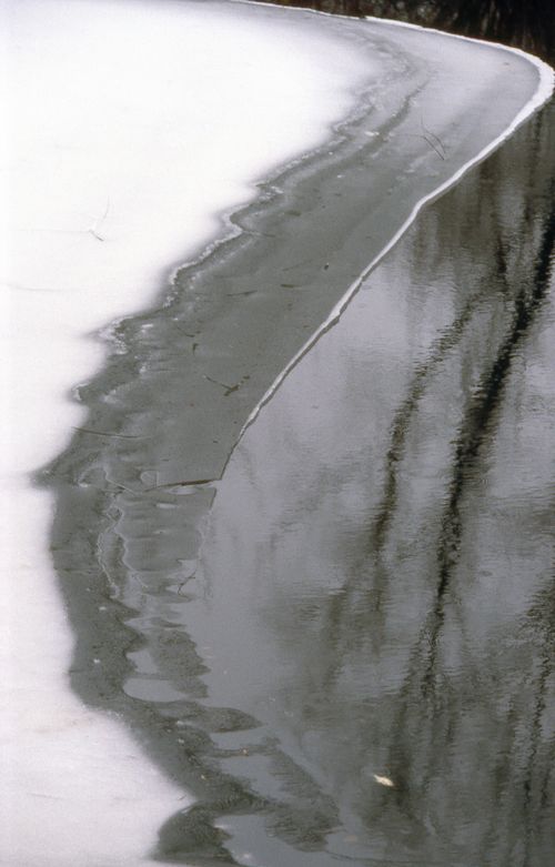 Free A serene winter scene of a partially frozen lake with snowy edges and tree reflections. Stock Photo