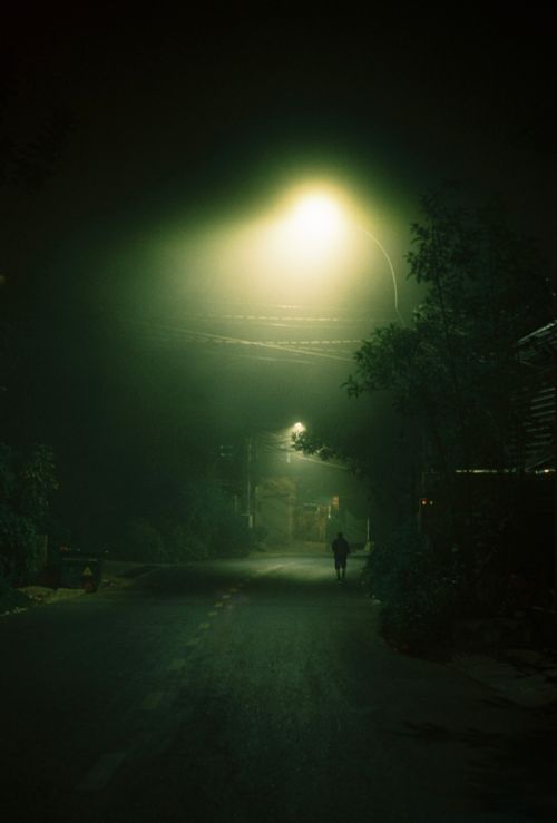 Free Eerie night photograph of a lone person walking under streetlights in a quiet village street. Stock Photo