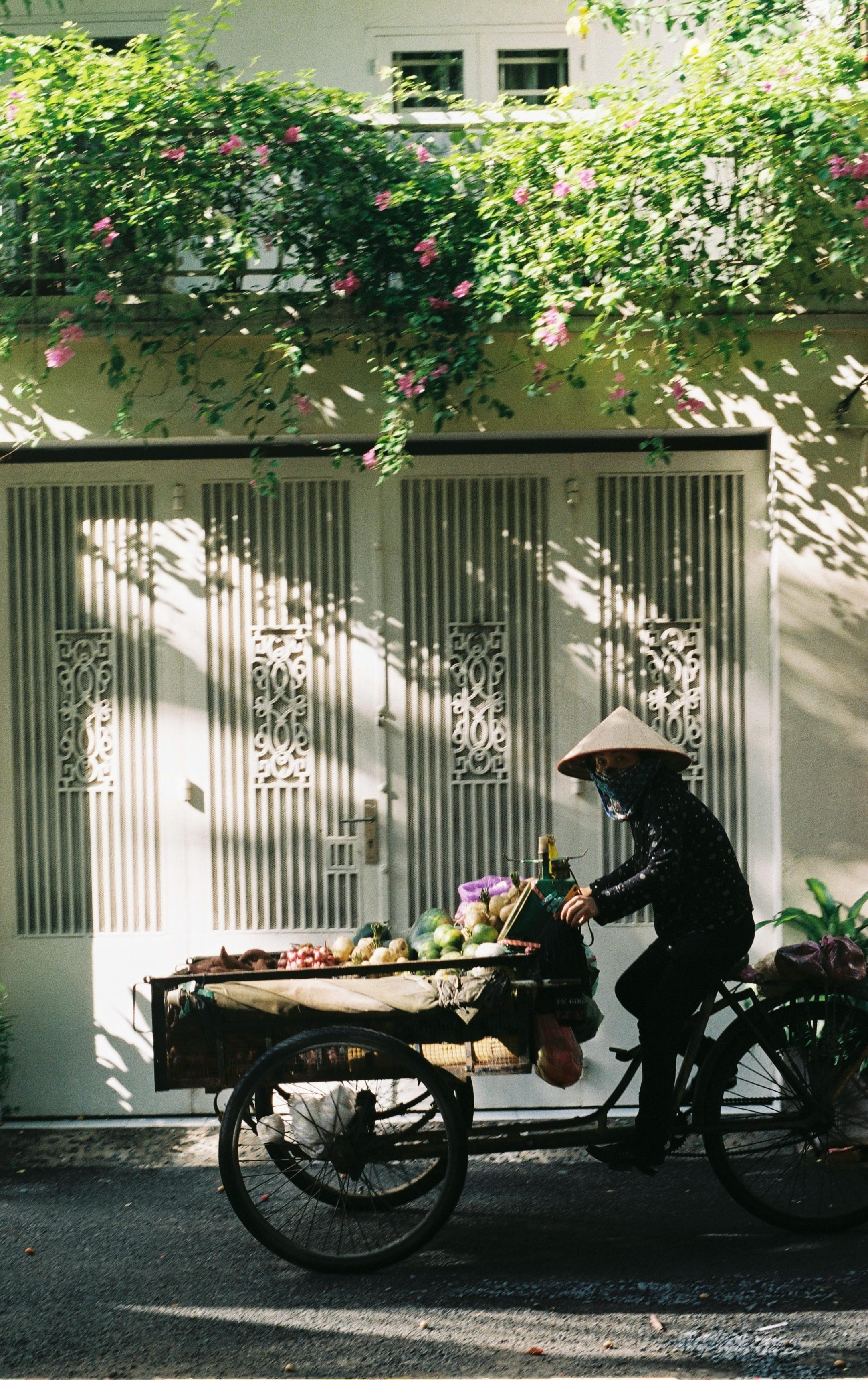 Asian man cycling with fruit cart in front of decorative building in Vietnam.