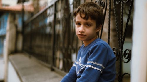 Free Thoughtful boy in blue sitting by an urban fence, selective focus. Stock Photo