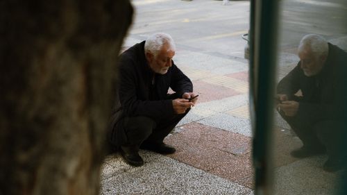 Free Elderly man in coat squatting on a sidewalk, focused on his smartphone. Stock Photo