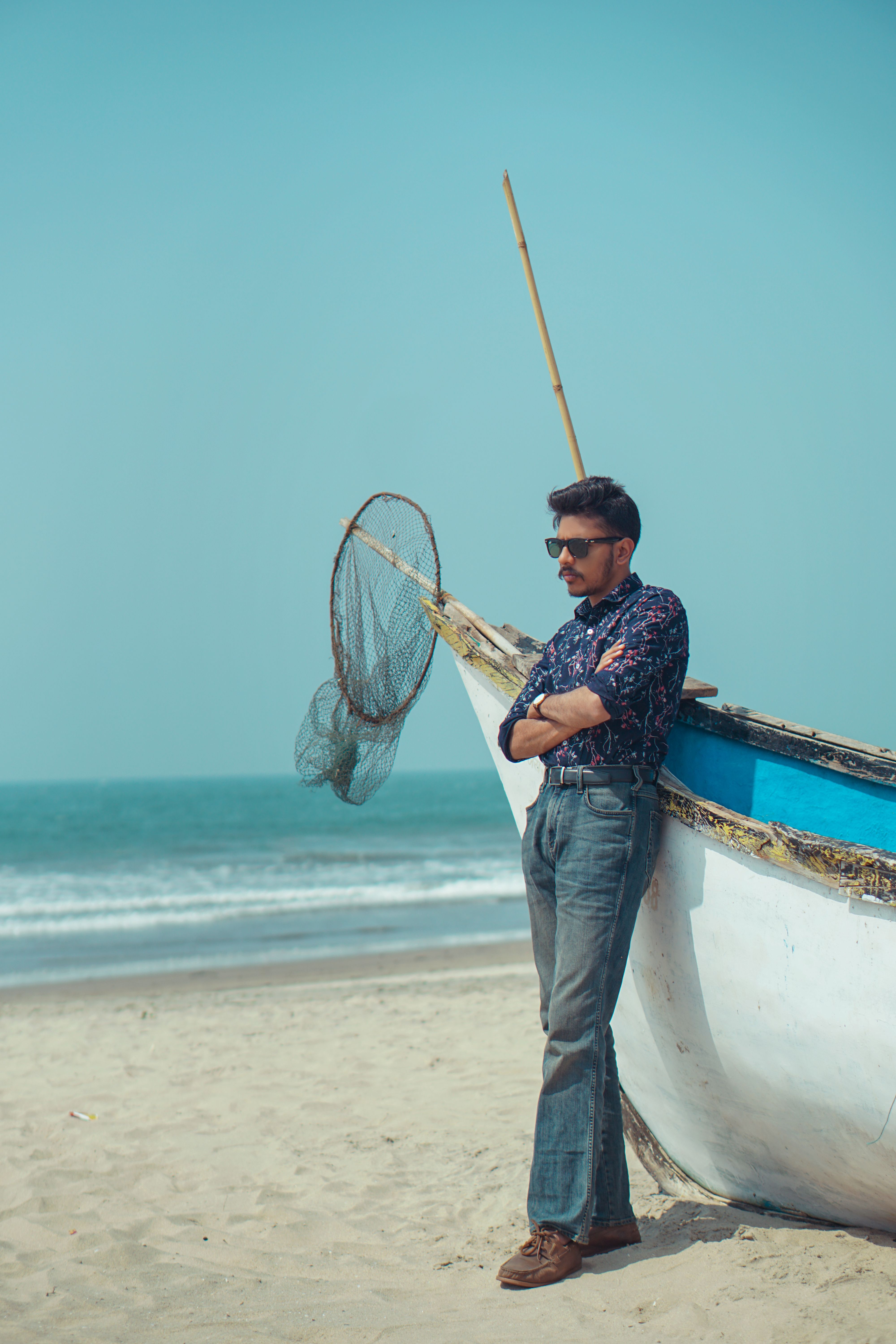 Man in sunglasses posing stylishly beside a boat on a sunny beach.