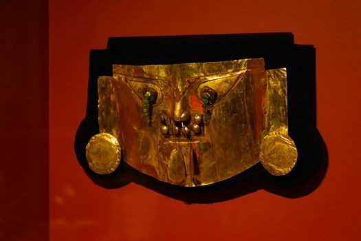 A golden artifact mask displayed on a vibrant red wall, showcasing intricate metalwork.
