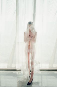Elegant woman posing in minimal lingerie behind sheer curtains, creating a soft and alluring atmosphere.