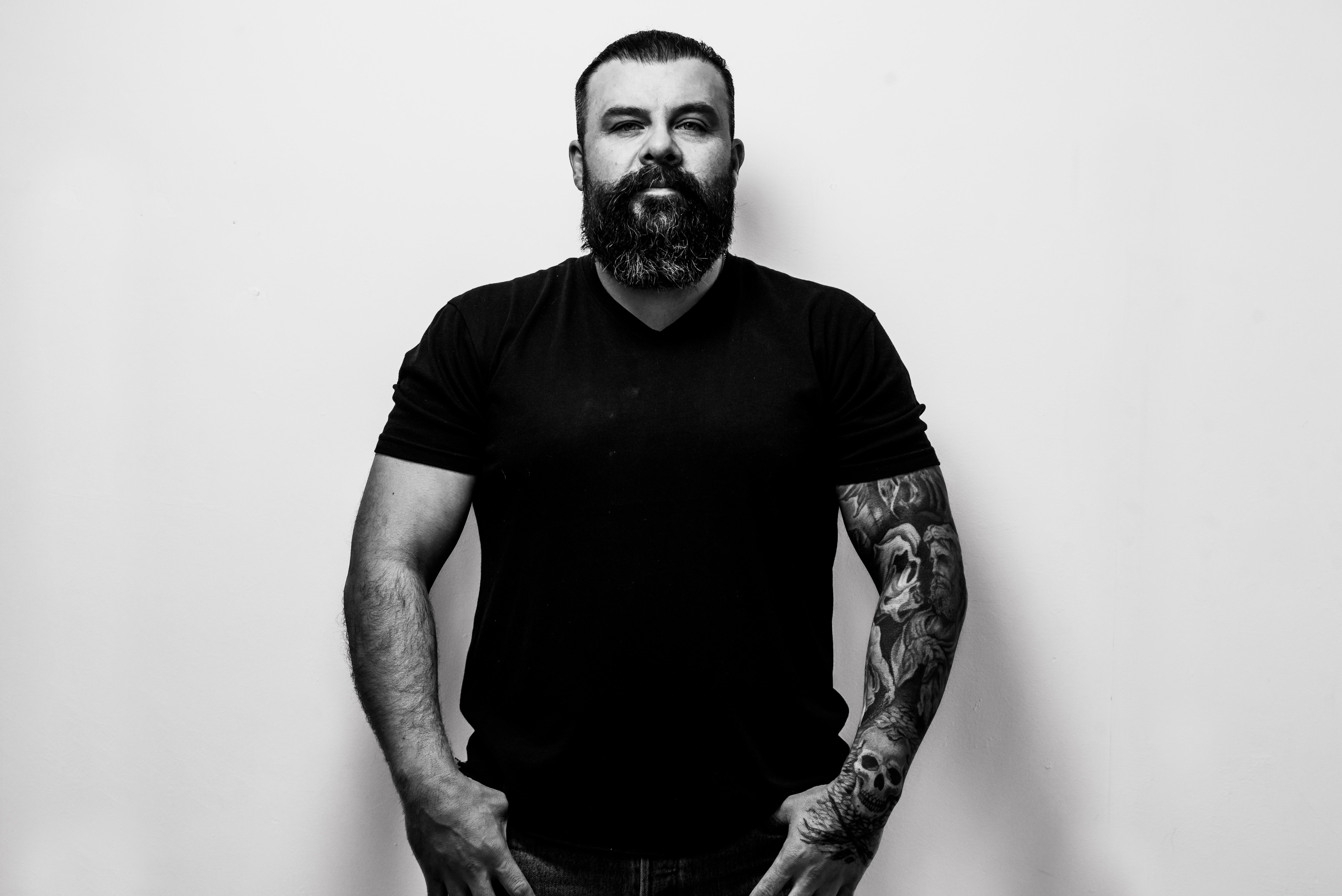 Portrait of a bearded man with tattoos in a black t-shirt, standing confidently against a white background.