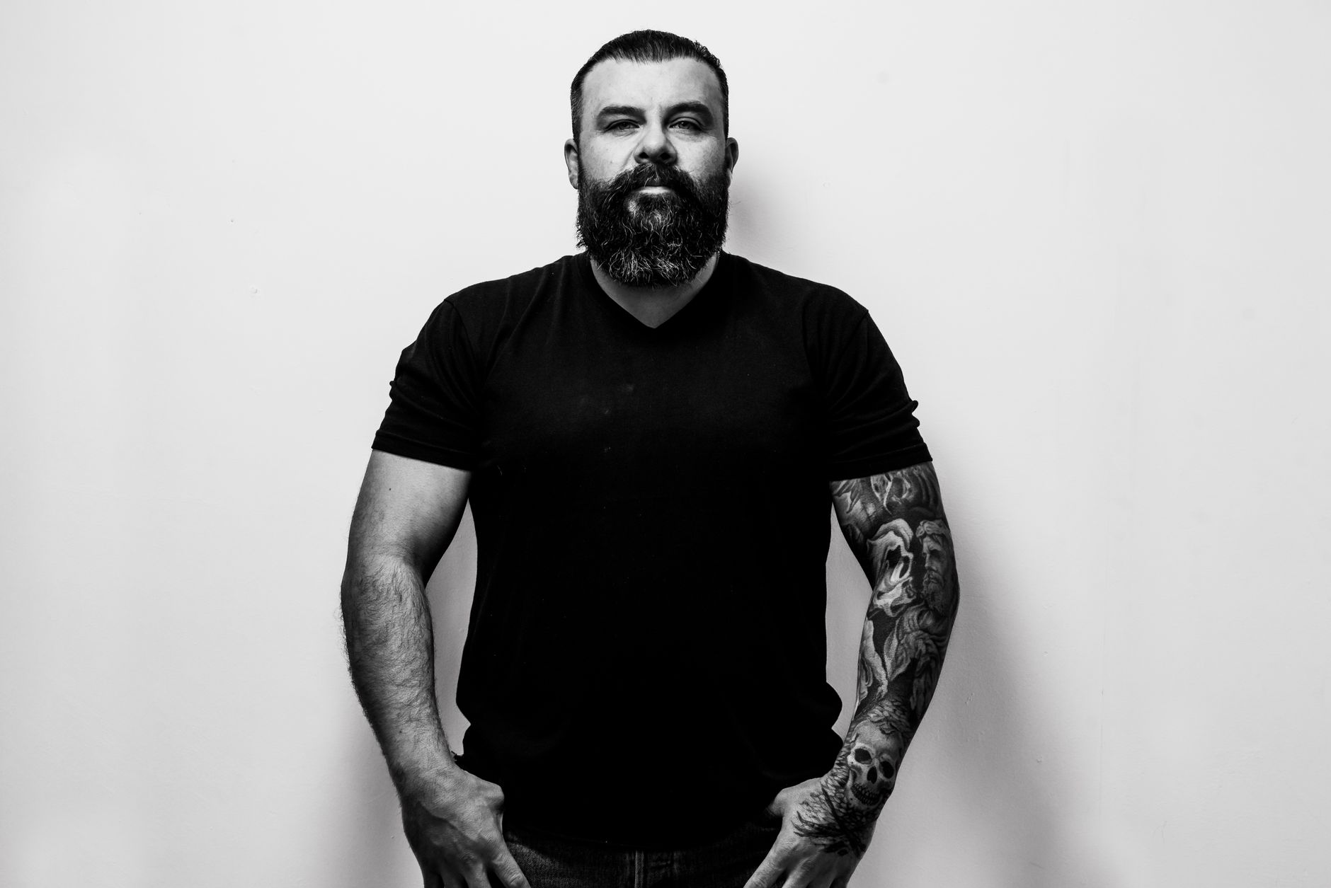 Portrait of a bearded man with tattoos in a black t-shirt, standing confidently against a white background.