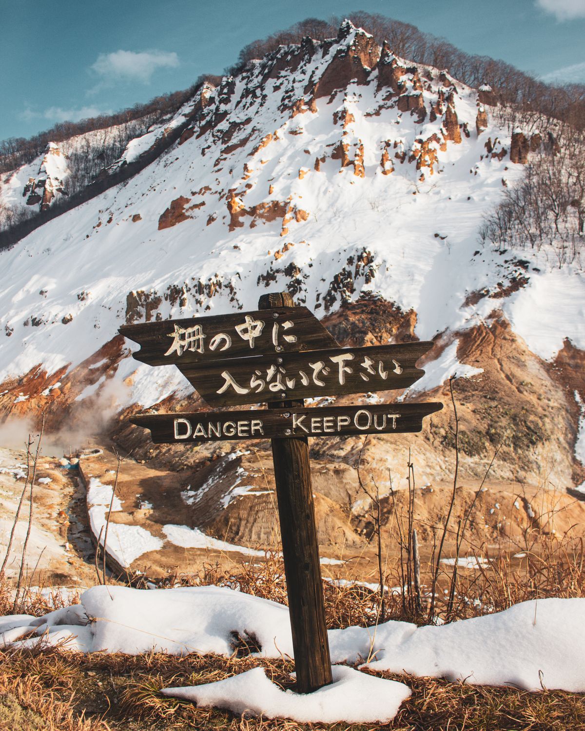 Wooden sign reading Danger Keep Out in Japanese and English beside a snowy volcanic slope in Hell Valley