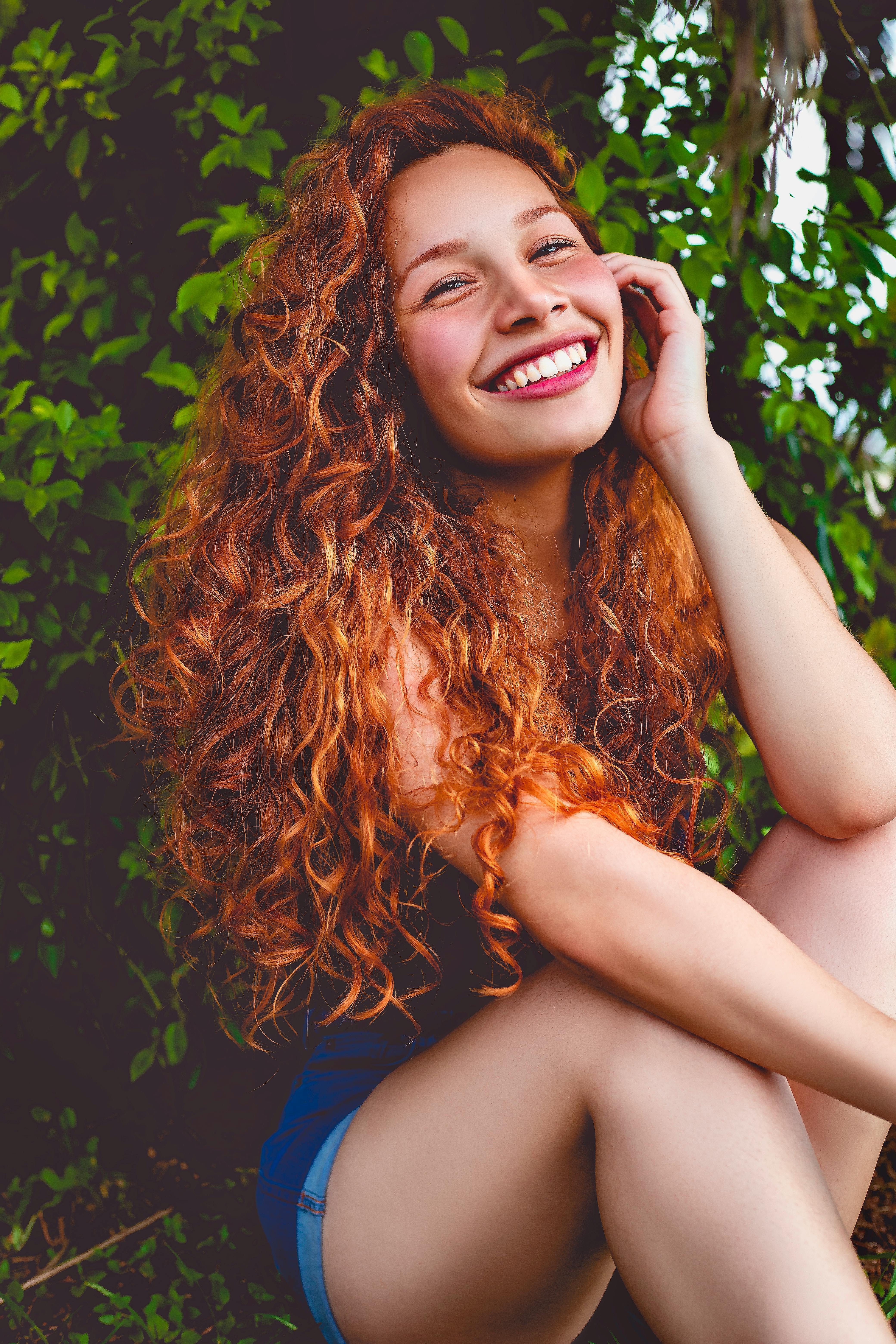 一位红发卷曲、面带微笑的女子在郁郁葱葱的户外环境中摆出姿势