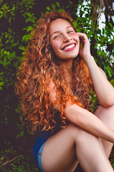 一位红发卷曲、面带微笑的女子在郁郁葱葱的户外环境中摆出姿势