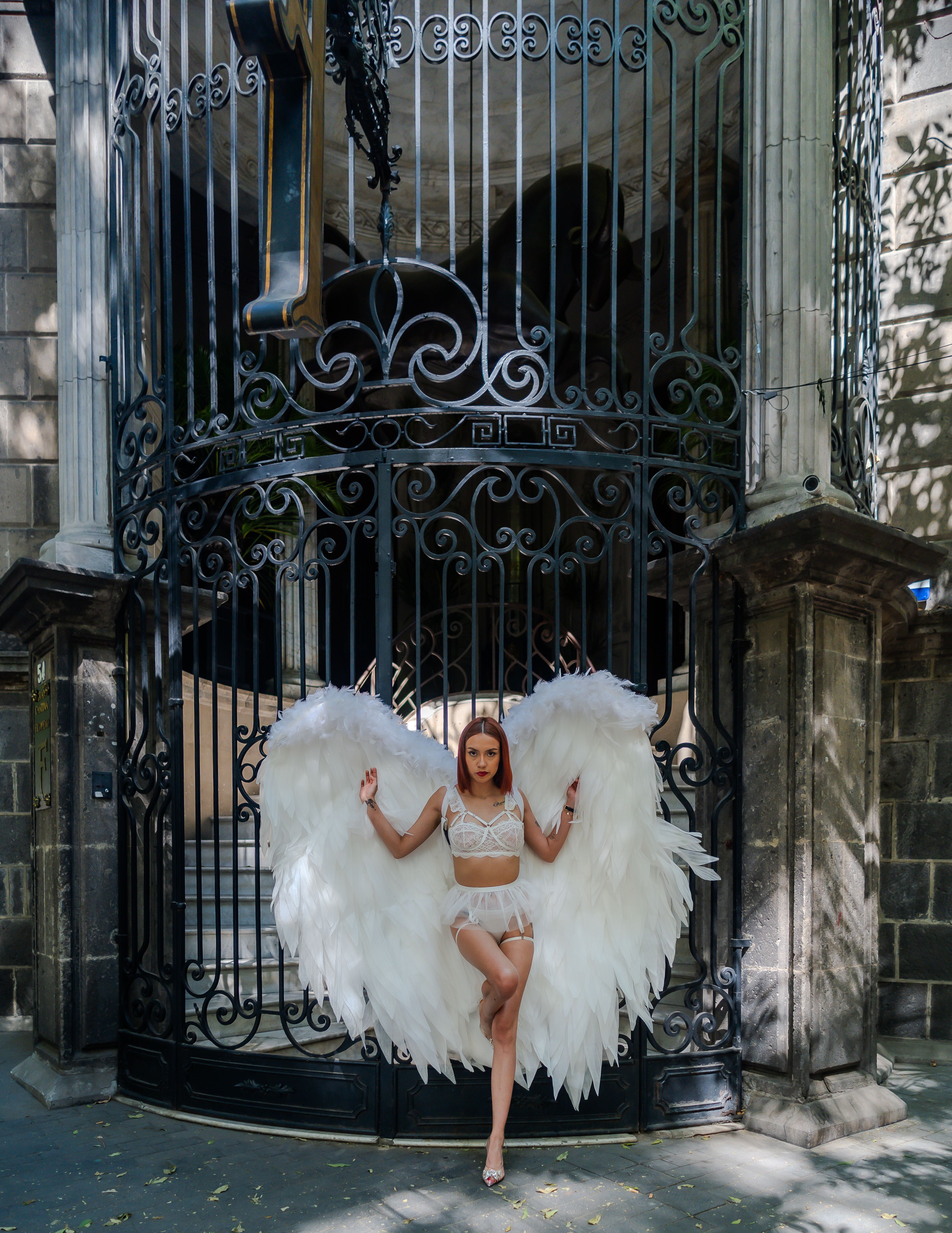 A woman in an angelic costume poses outdoors in front of a historic building gate in Mexico City.