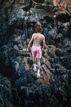 A shirtless man in pink shorts climbs a rocky terrain, showcasing courage and adventure.