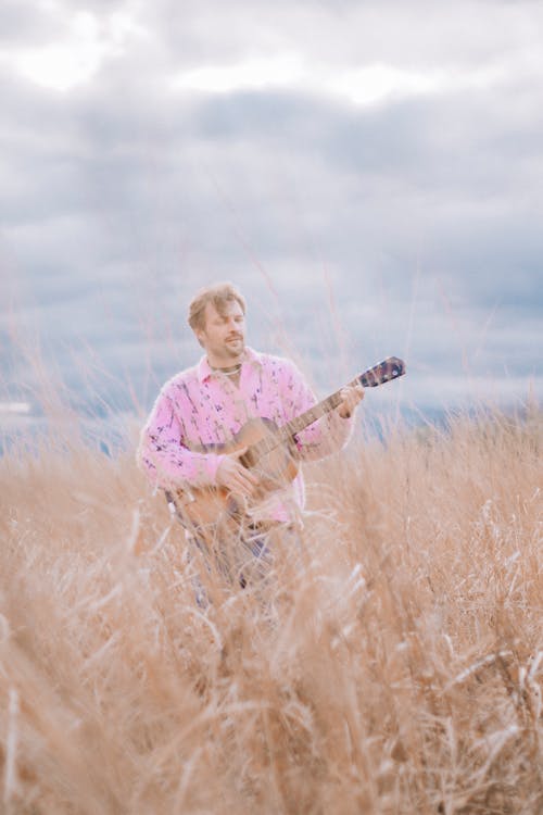 A man in a pink shirt playing an acoustic guitar in a field