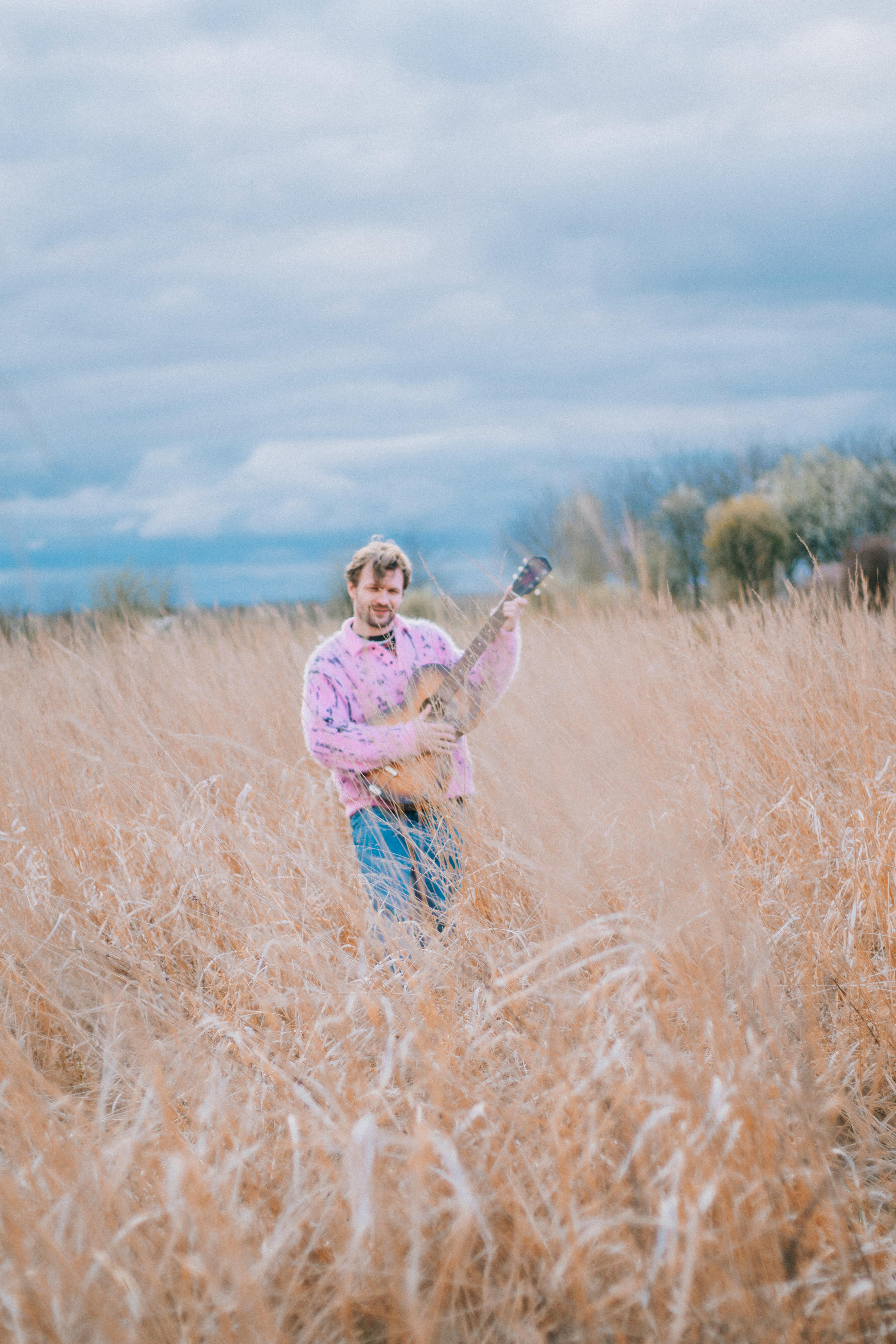 Man in a field playing an acoustic guitar under a cloudy sky, showcasing musical passion.