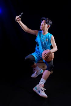 A teenager in sportswear takes a selfie while holding a basketball, indoors with dramatic lighting.