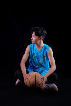 Teen boy in blue sportswear sitting with basketball on a black background.