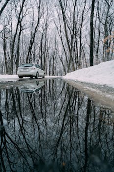 白雪皑皑的道路上，一辆白色汽车驶过，水坑中倒映着树木，捕捉到了宁静的冬日景象