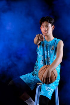 A young basketball player in a blue uniform sits on a stool in a studio setting, holding a basketball.