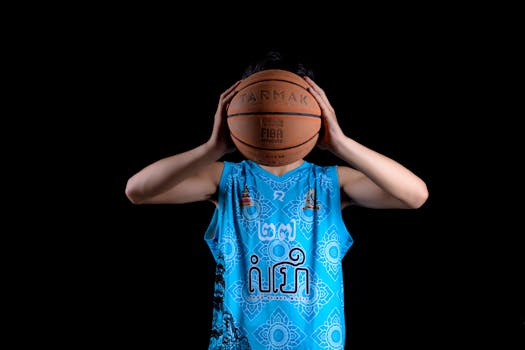 A teenager holds a basketball in front of his face, wearing a detailed blue jersey.