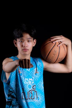 Asian teenager in blue tank top holding a basketball against a black background, showcasing confidence and sportsmanship.