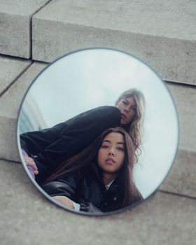 Artistic photo of two women reflected in a circular mirror on urban stairs, showcasing fashion.