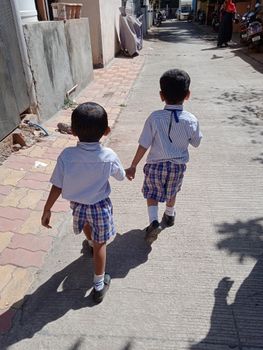 Two young schoolchildren in uniforms walking hand in hand along a sunlit street.